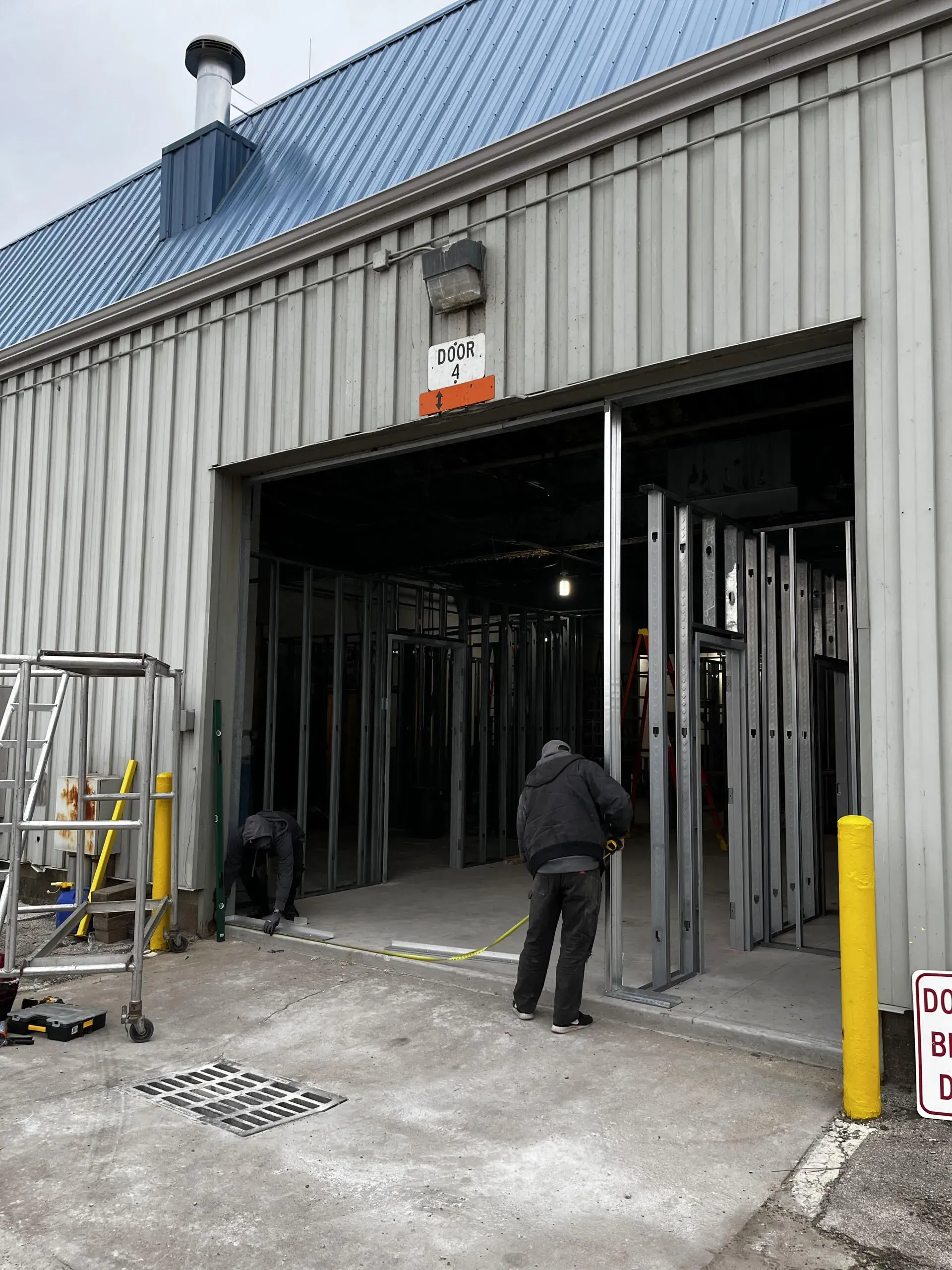 A worker installs metal framing inside a building entrance, while another worker kneels in the background on a grey lot.