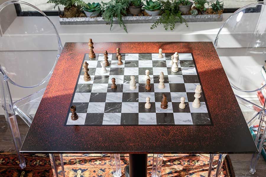 A chess set with pieces scattered on a dark, speckled table between two transparent chairs near a window with plants.