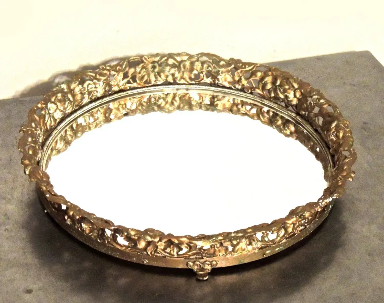 A round, vintage-style vanity tray with a mirrored base and a decorative, ornate gold-toned metal filigree border.