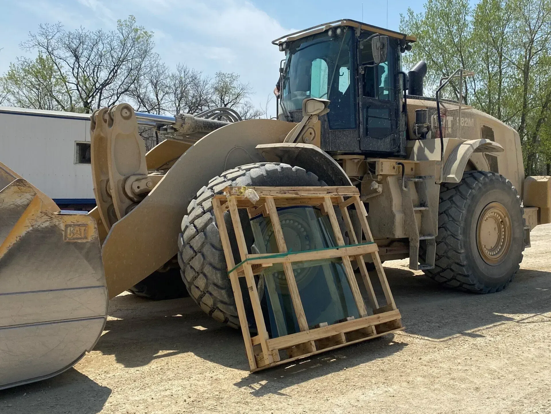 A large, tan Caterpillar wheel loader parked on gravel with a wooden crate holding a glass pane resting against its tire.