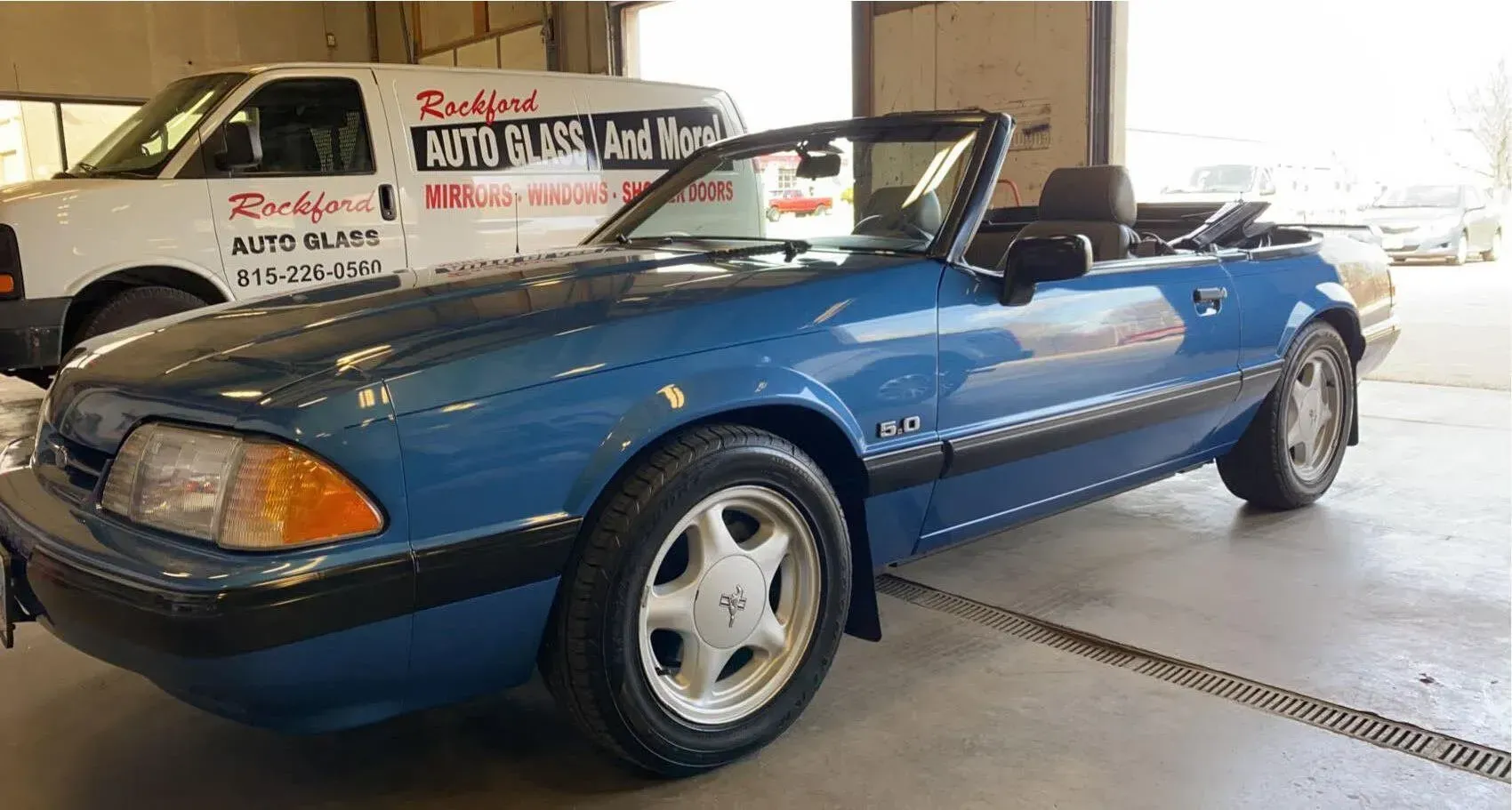 A blue Ford Mustang convertible parked in an auto repair shop next to a work van.
