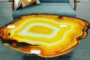 A circular coffee table with a polished yellow and brown agate slice top, set in front of a blue sofa.