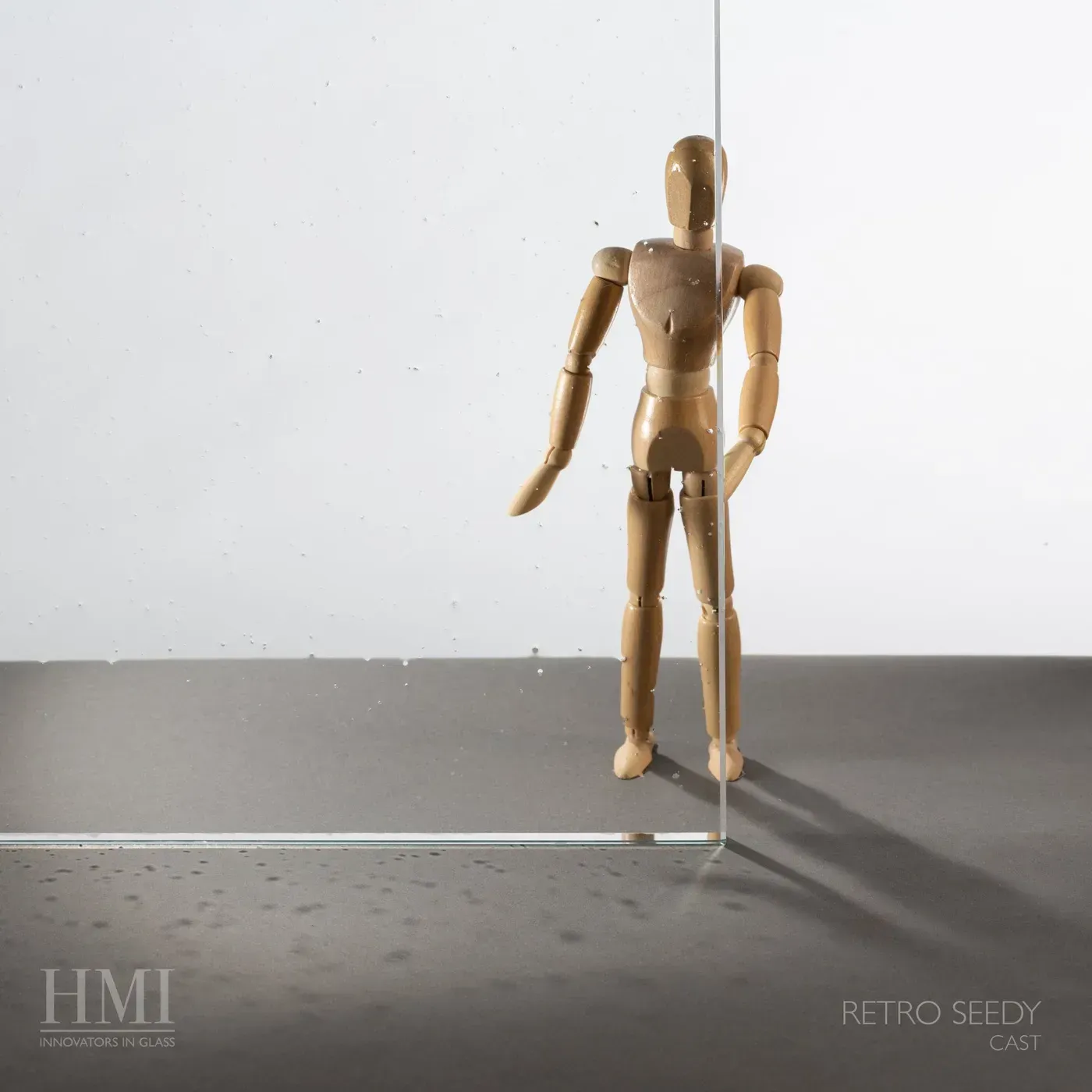 A wooden artist’s mannequin stands behind a transparent glass panel, casting a shadow on a dark surface. HMI logo present.