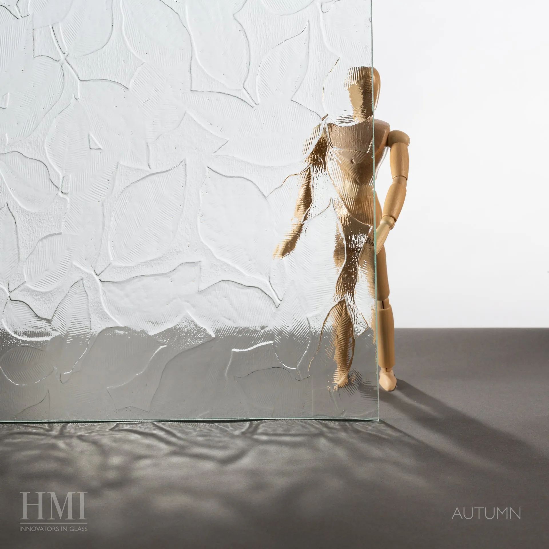 A wooden mannequin stands behind a sheet of textured Autumn-patterned glass, casting a distorted shadow on the surface.