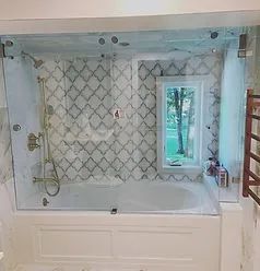 A modern bathroom with a white bathtub, a brass shower fixture, and a window against a patterned tile wall.