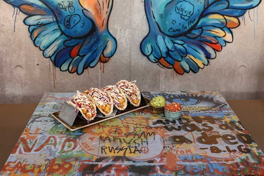 Four tacos in a metal holder with two small bowls of salsa, set on a colorful graffiti-covered table against wall art.