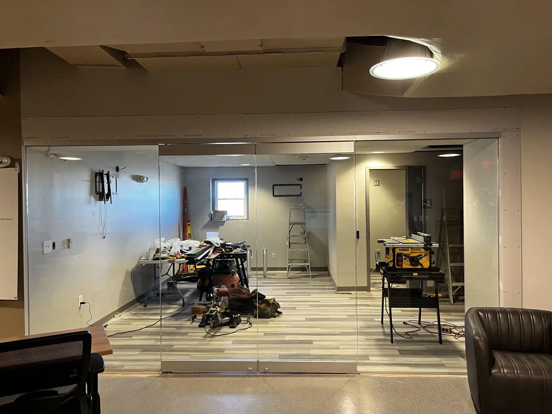 A glass-walled room under construction, with tools and equipment inside, seen from an adjacent office space.