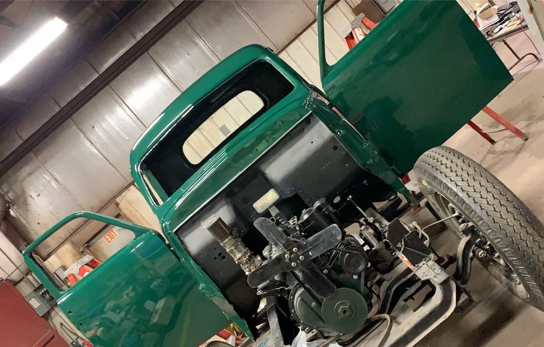 A green vintage truck cab under restoration, featuring an exposed engine and single front tire in a garage setting.