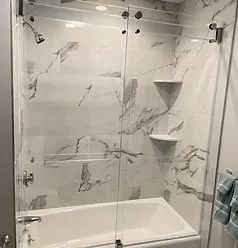 A glass sliding shower door with black hardware installed over a white bathtub against a white marble-patterned wall.