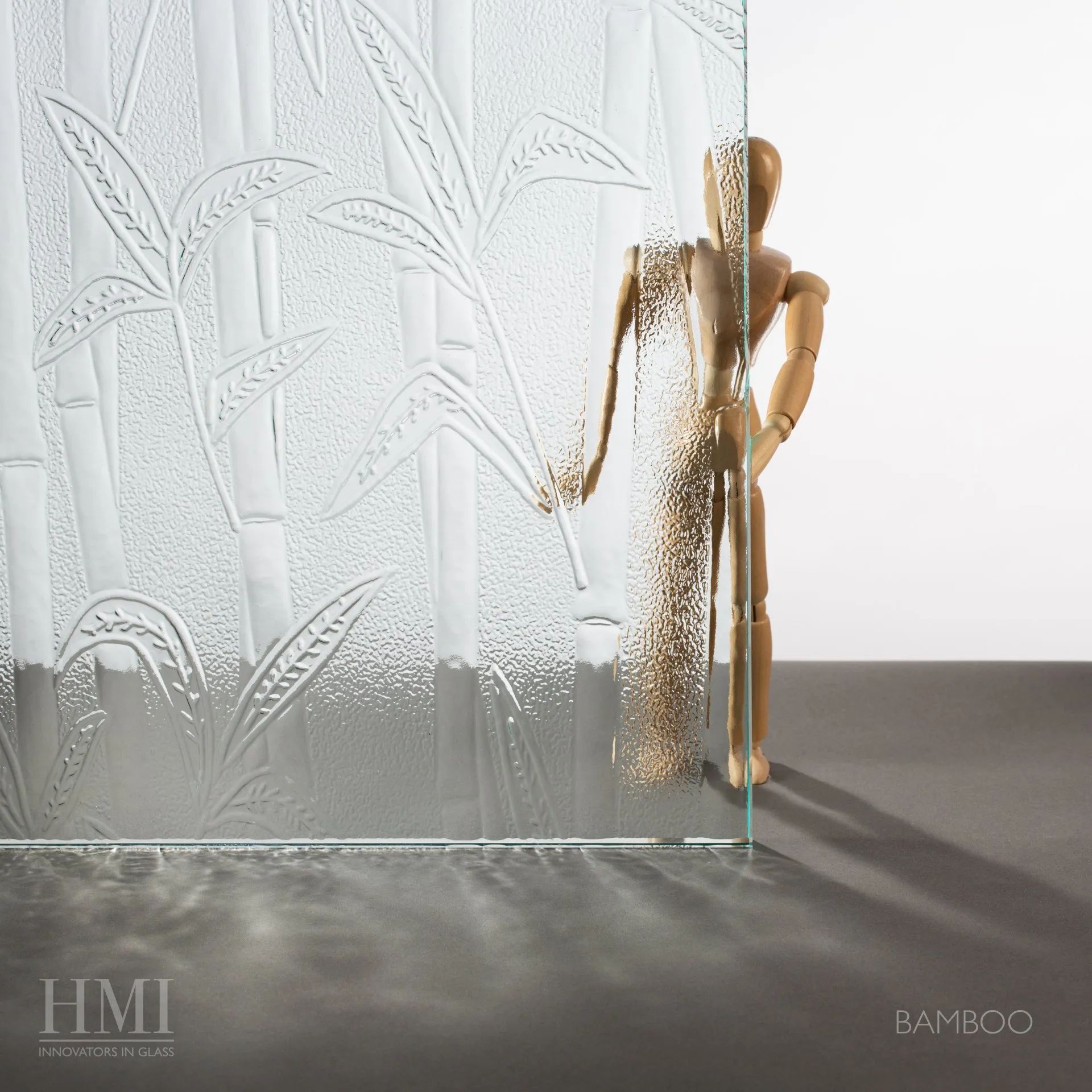 A wooden mannequin stands behind a patterned glass panel embossed with bamboo designs, casting a shadow on a dark surface.