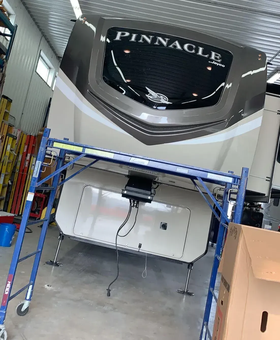 A Pinnacle fifth-wheel camper parked indoors, partially obscured by a blue metal scaffolding frame.