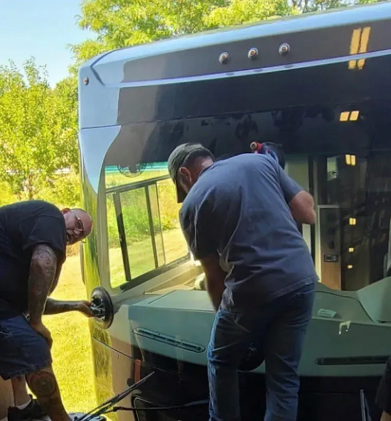 Two workers using suction cups to install a large windshield onto the front of a bus outdoors.