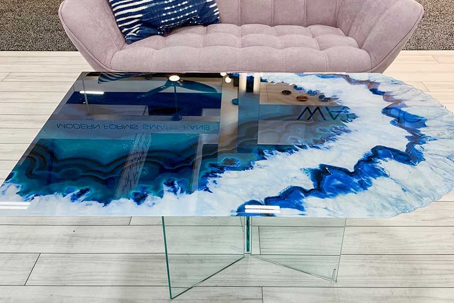 A modern coffee table with an agate-inspired blue and white resin tabletop, resting on clear glass legs before a sofa.