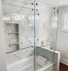 A modern bathroom featuring a white marble-tiled bathtub area with a glass sliding door and a built-in recessed shelf.