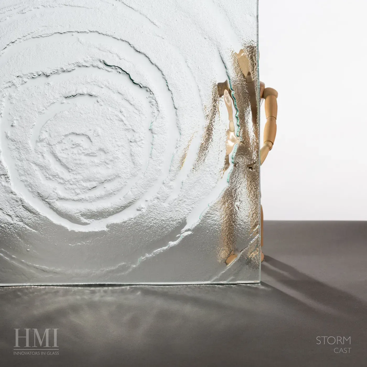 A wooden artist's mannequin stands behind a textured glass panel featuring a concentric, ripple-like pattern.