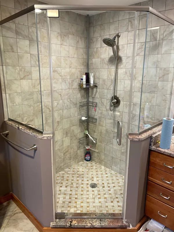 A corner walk-in shower with tiled walls, glass panels, a metal showerhead, and corner shelving, in a neutral bathroom.