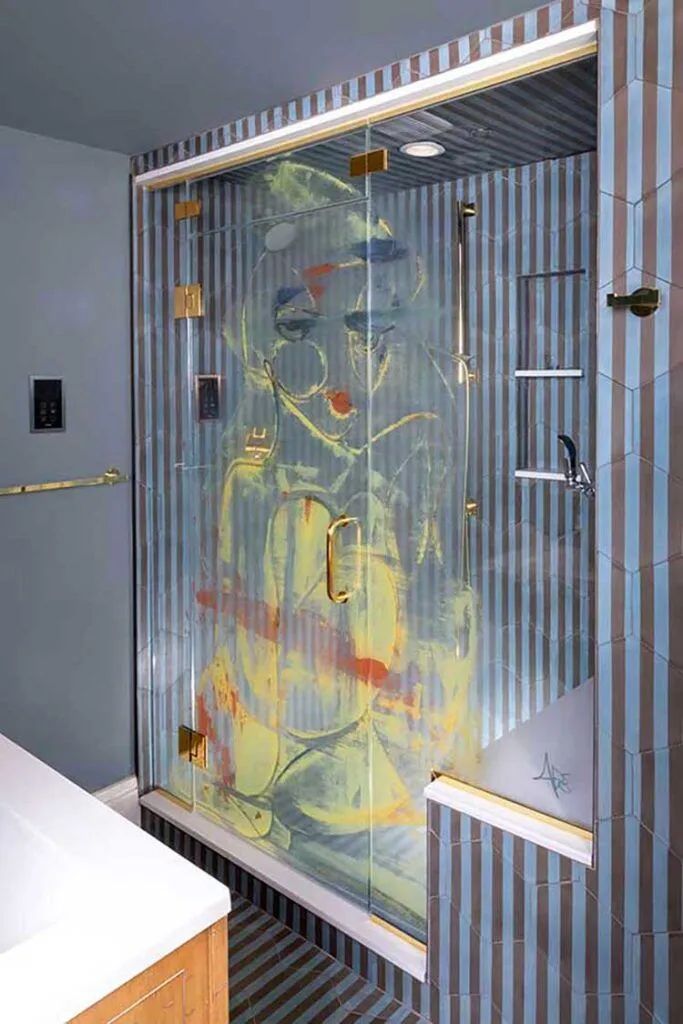 A walk-in shower with a glass door featuring a large, abstract colorful figure, set against vertical blue-grey tiles.