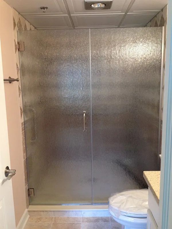 A glass-enclosed shower with textured privacy glass, installed in a bathroom next to a toilet.