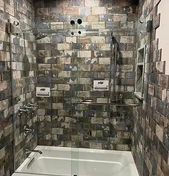 A white bathtub with a clear glass door enclosure against a rustic, multi-colored brick-tiled wall in a shower.