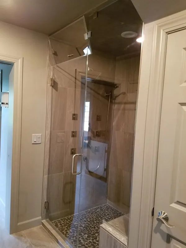 A frameless glass shower enclosure with a chrome handle, tiled walls, and pebble-style flooring in a bathroom.