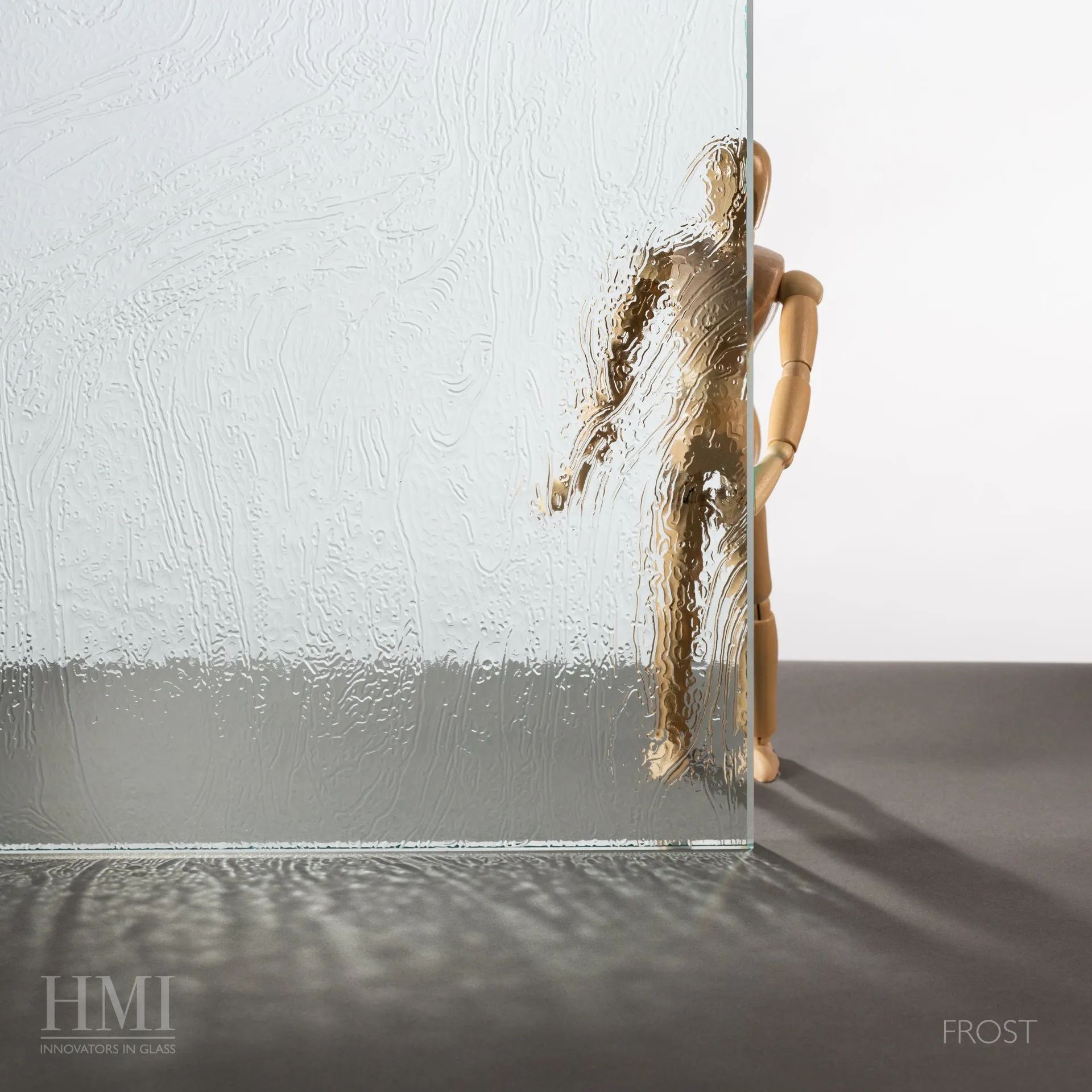 A wooden mannequin stands behind a sheet of textured, translucent glass, which distorts the view of the figure.