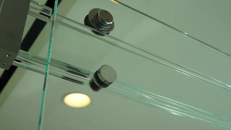 Close-up of a glass shower door with metal rollers attached to a top track, set against a white wall with a ceiling light.