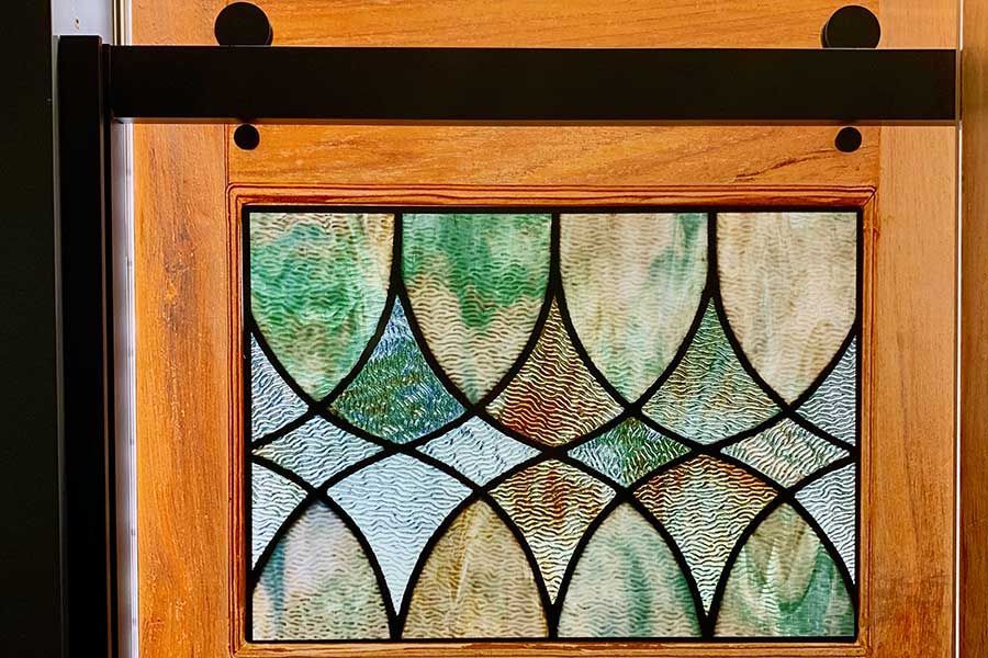 A stained glass window featuring a green, white, and brown repeating arch pattern, set within a wood frame with a black rail.