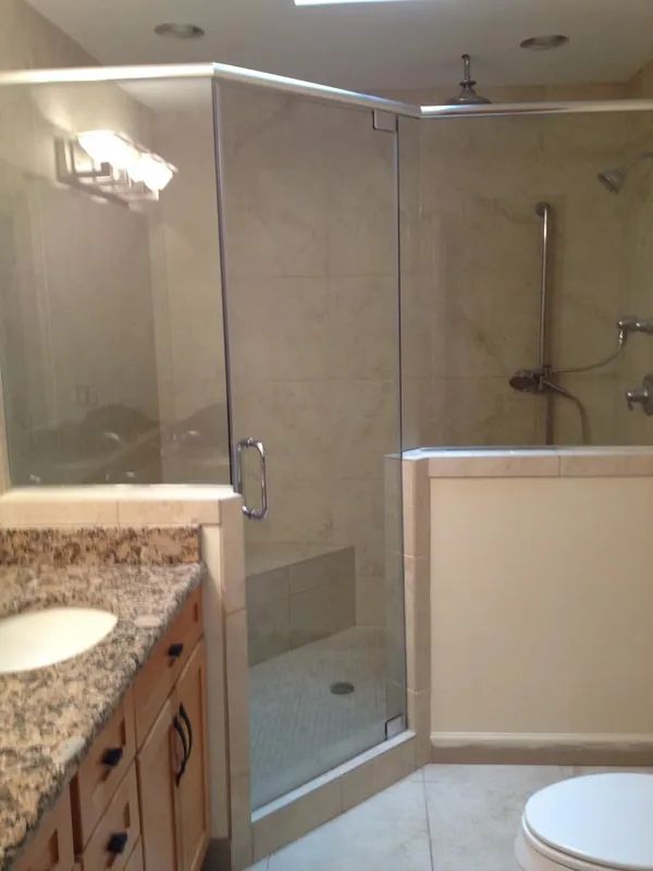 A glass-enclosed corner shower with a granite vanity countertop and light-colored tile flooring in a modern bathroom.