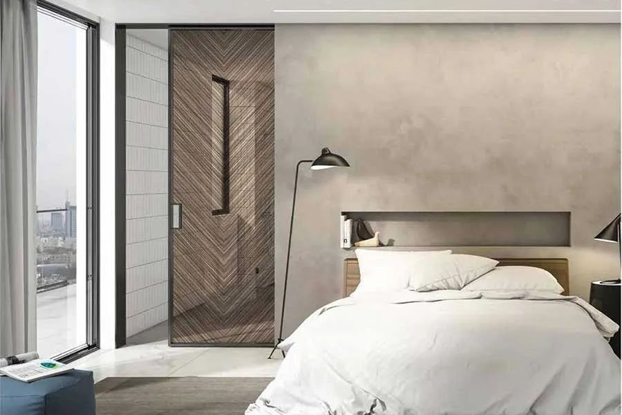 A modern bedroom with a herringbone-patterned wooden sliding door, a white bed, and a floor lamp against a beige wall.