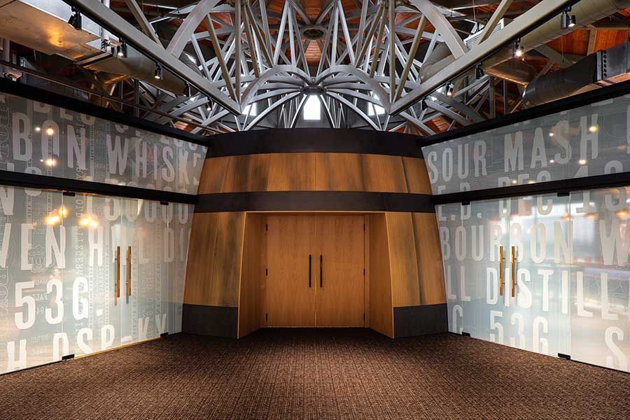 Interior of a distillery visitor center with a wooden barrel-shaped entrance flanked by frosted glass walls featuring text.