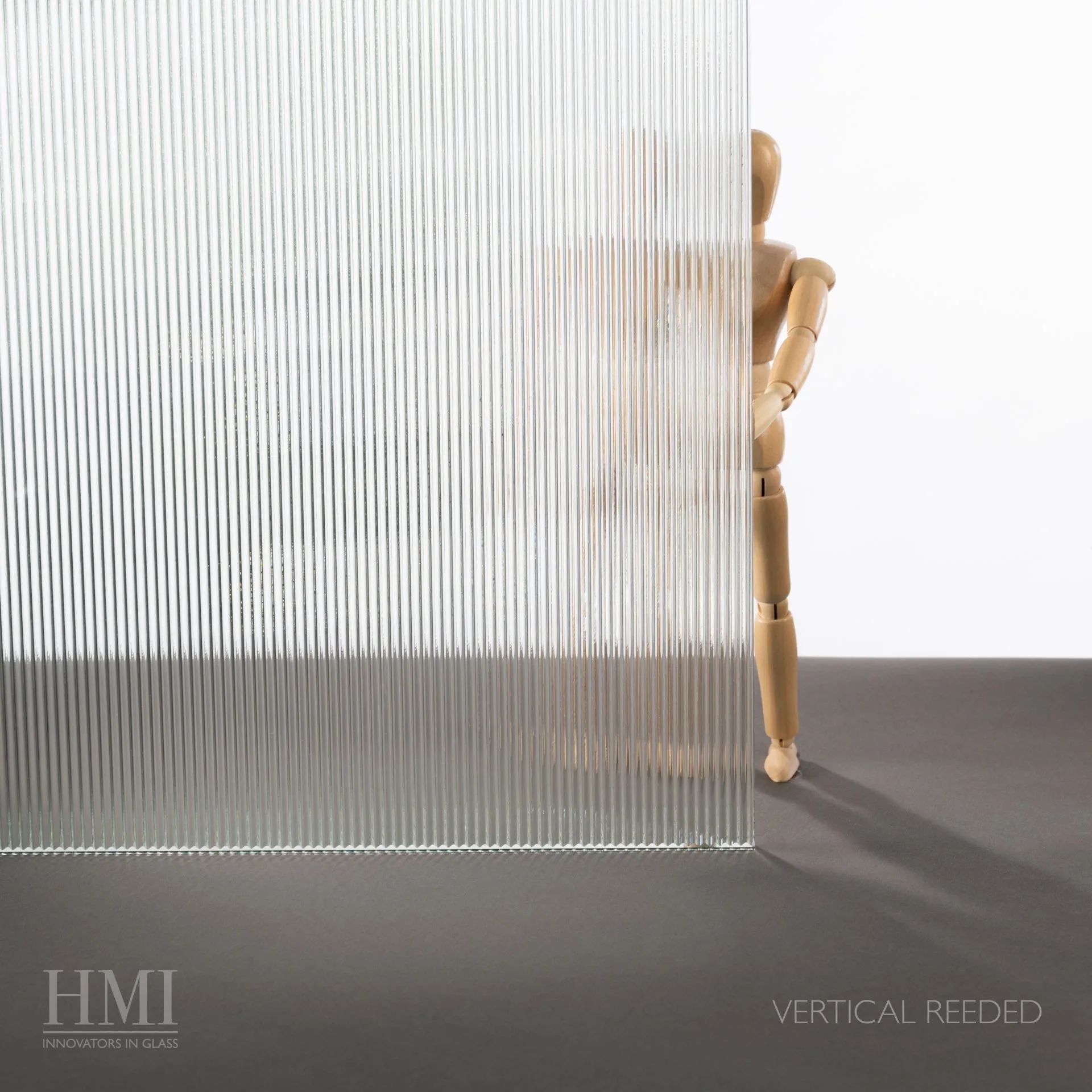 A sheet of vertical reeded glass obscuring a wooden artist mannequin on a dark surface, with HMI logo text in the corner.
