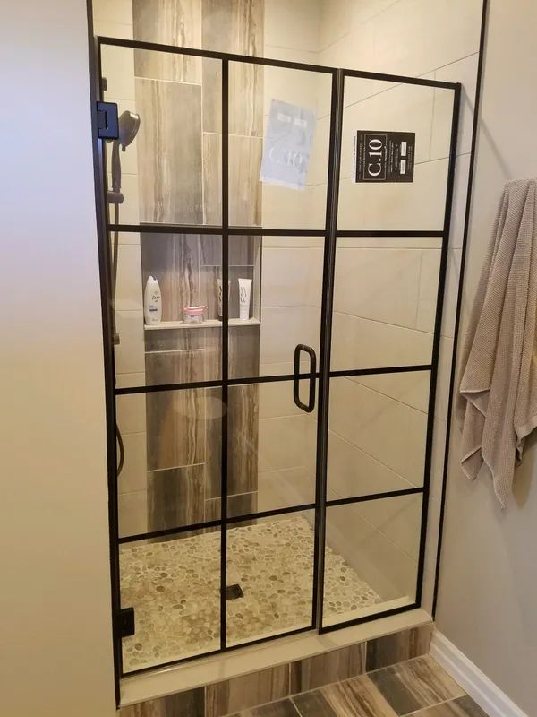 A tiled walk-in shower with a black-framed grid glass door and pebble flooring, featuring a built-in shampoo niche.