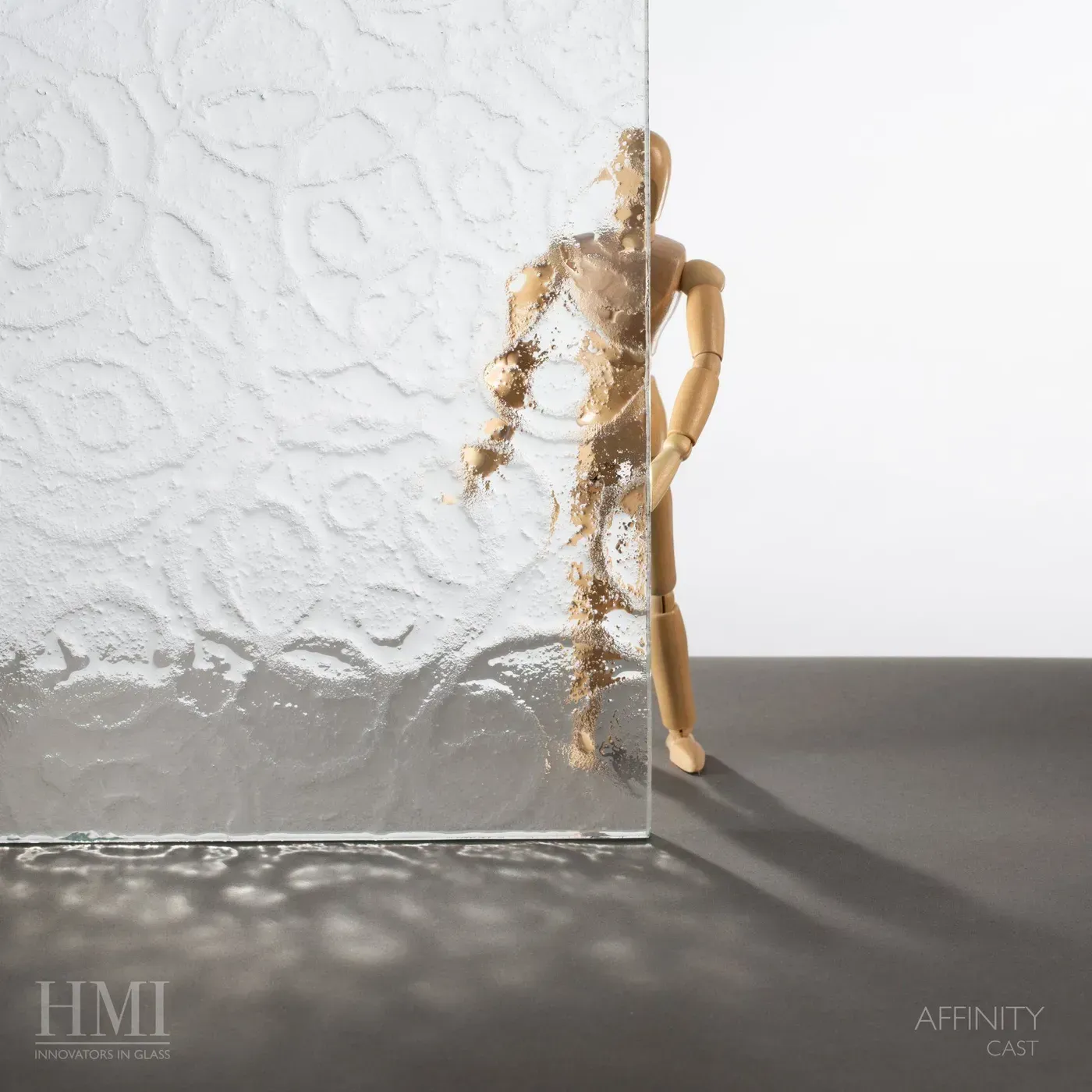A wooden artist’s mannequin stands behind a clear, textured glass panel that distorts its form, casting dappled shadows.