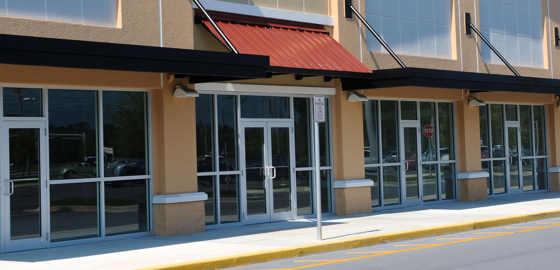 A row of vacant storefronts with glass doors, large windows, tan stucco walls, and black and red fabric awnings.