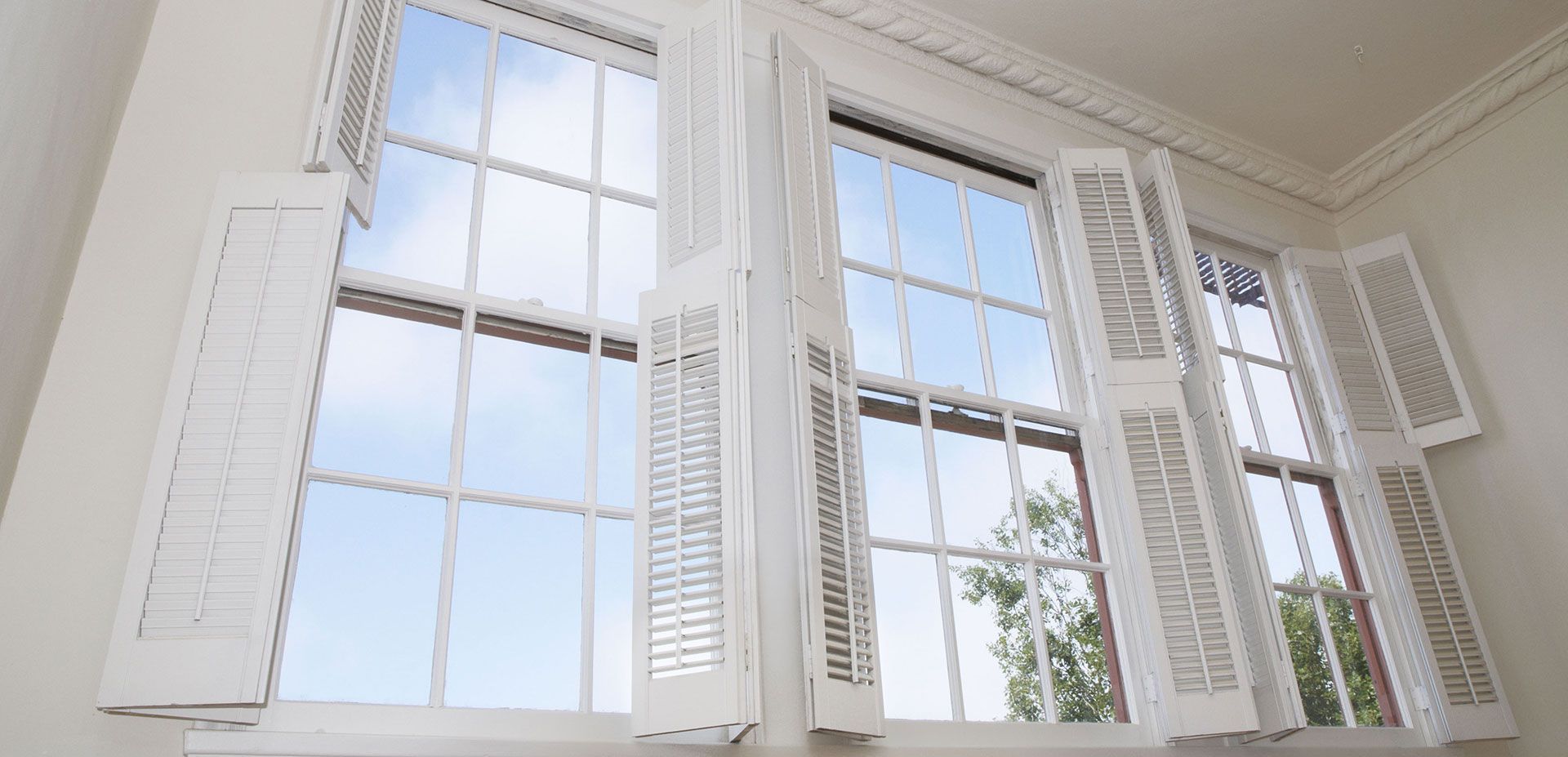 White plantation shutters are partially opened over three large, multi-pane windows looking out at a bright sky.