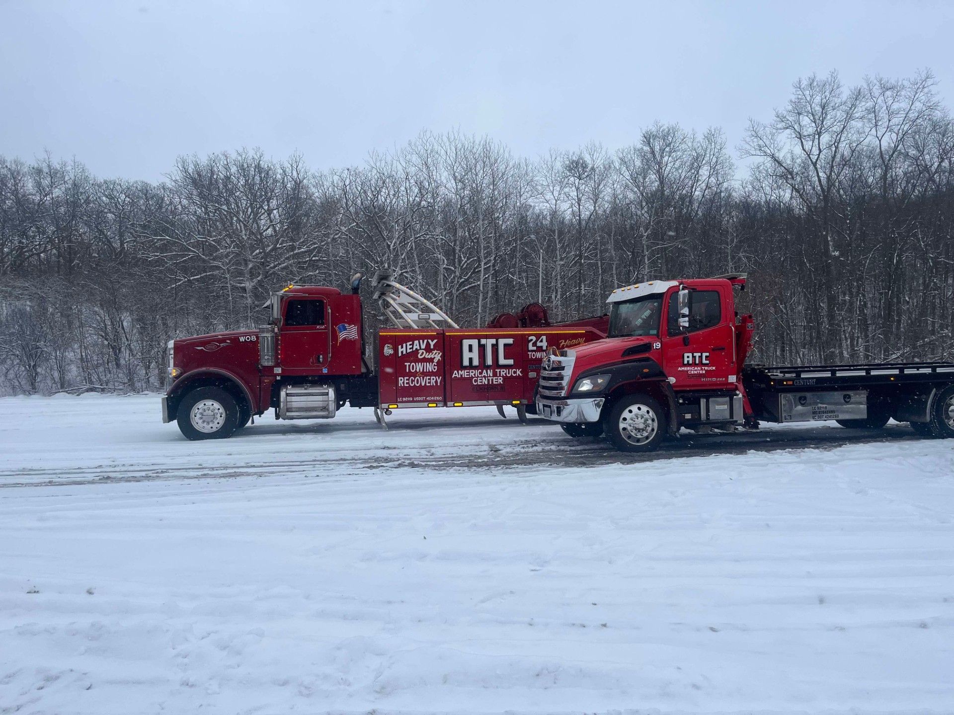 Two red tow trucks on a snow-covered field with trees in the background.