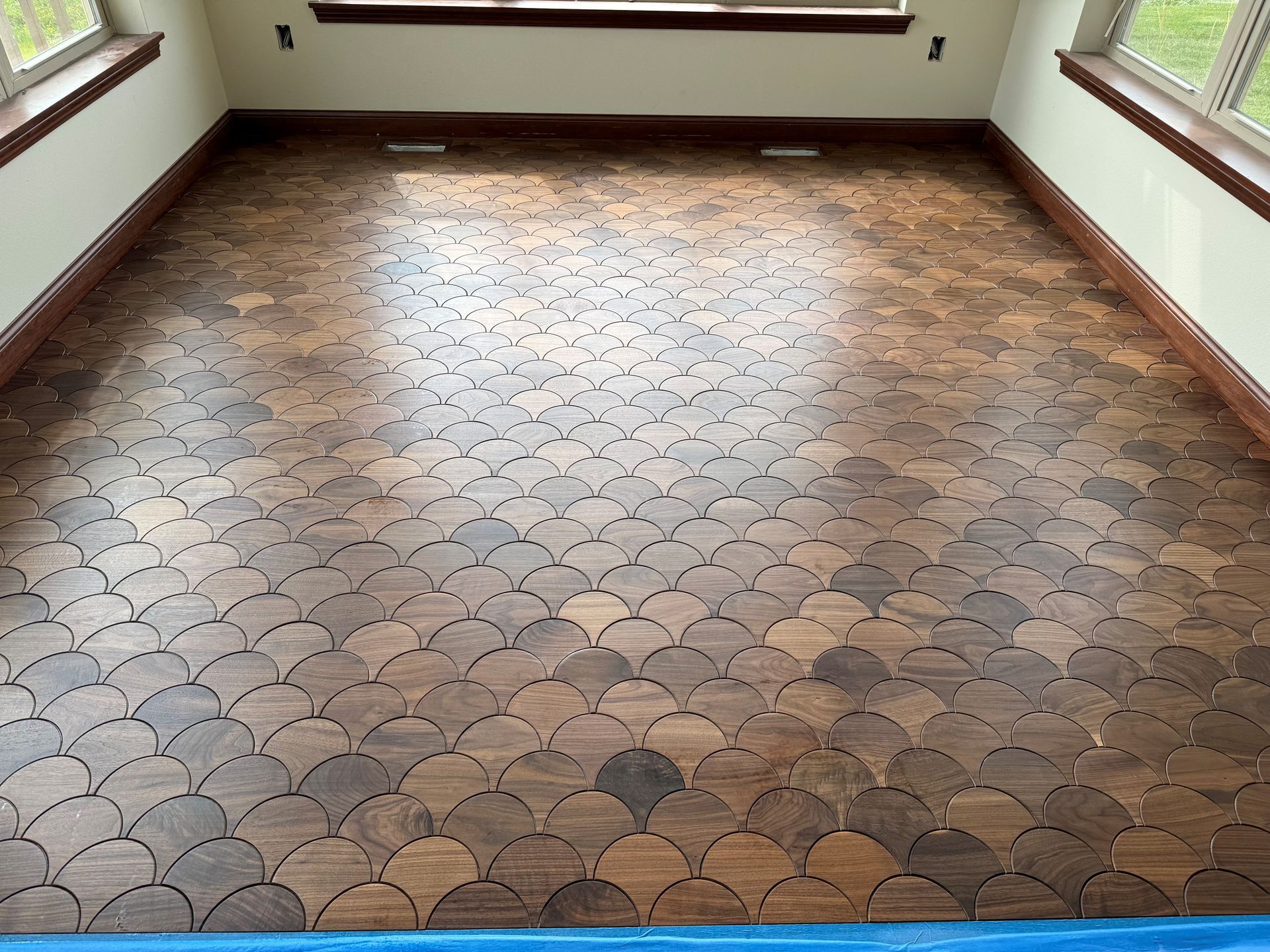 A dark brown, scallop-patterned wooden floor fills a sunlit room, surrounded by light walls and windows.