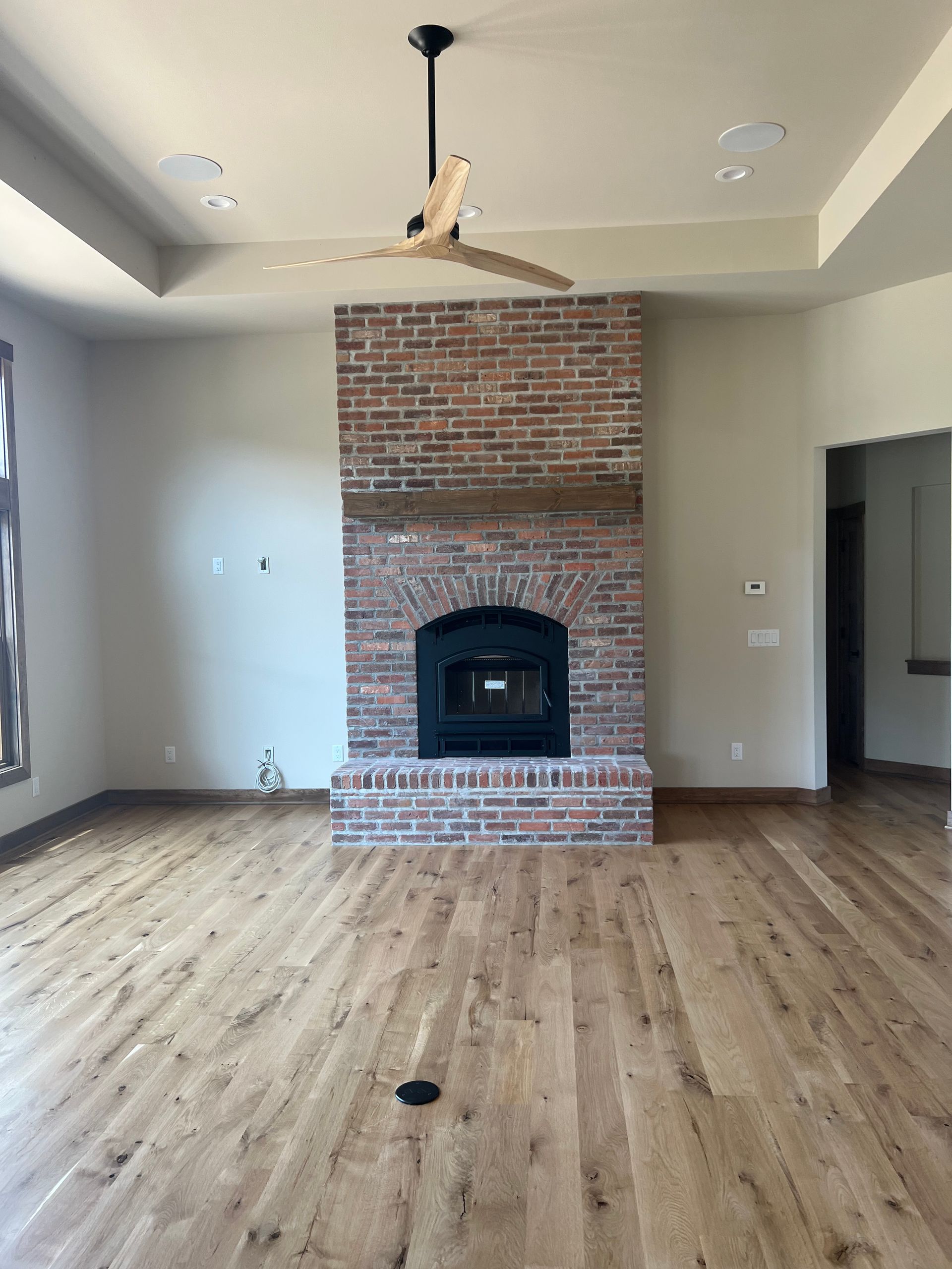Brick fireplace with black insert and wooden mantle in a room with hardwood floors and a ceiling fan.