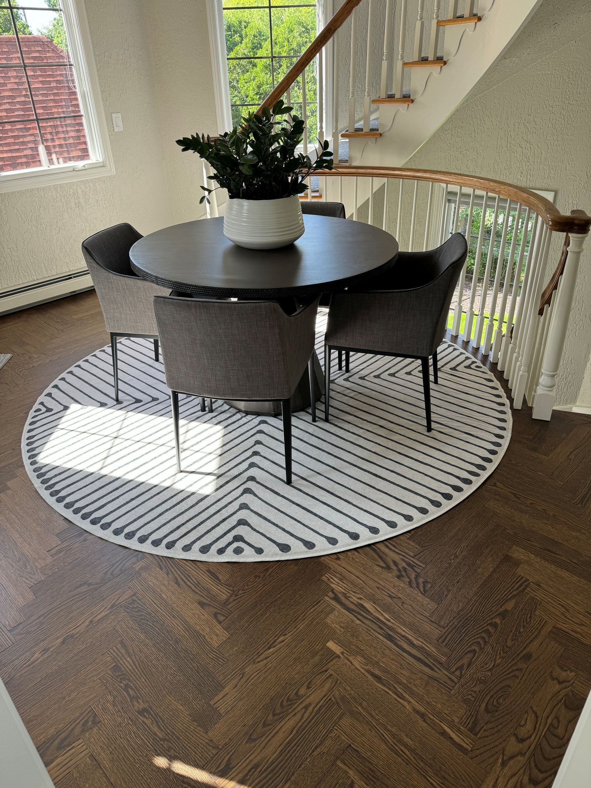 Round dining table with four chairs on a patterned rug, by a staircase, set on a wood floor.