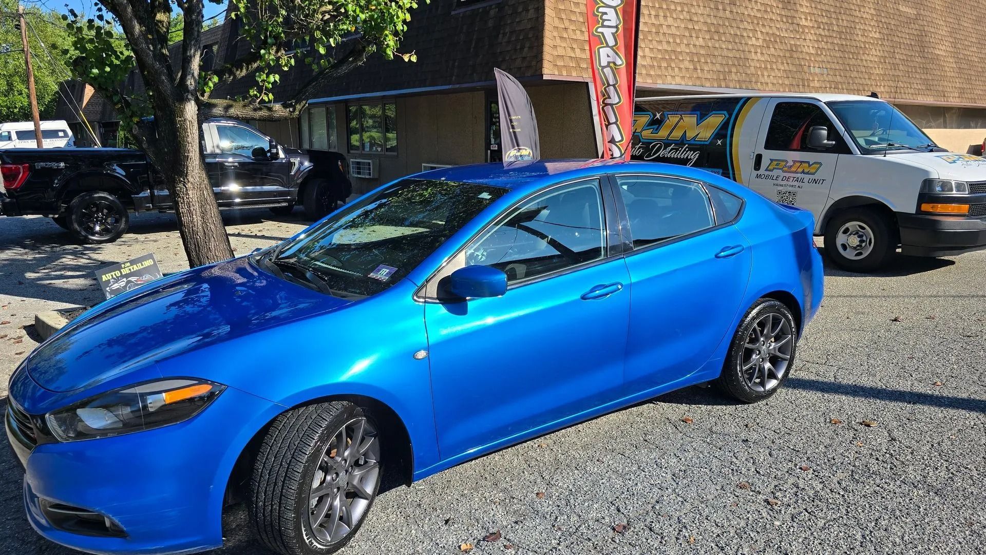 A bright blue Dodge Dart sedan parked on a gravel lot in front of a commercial building with other vehicles nearby.