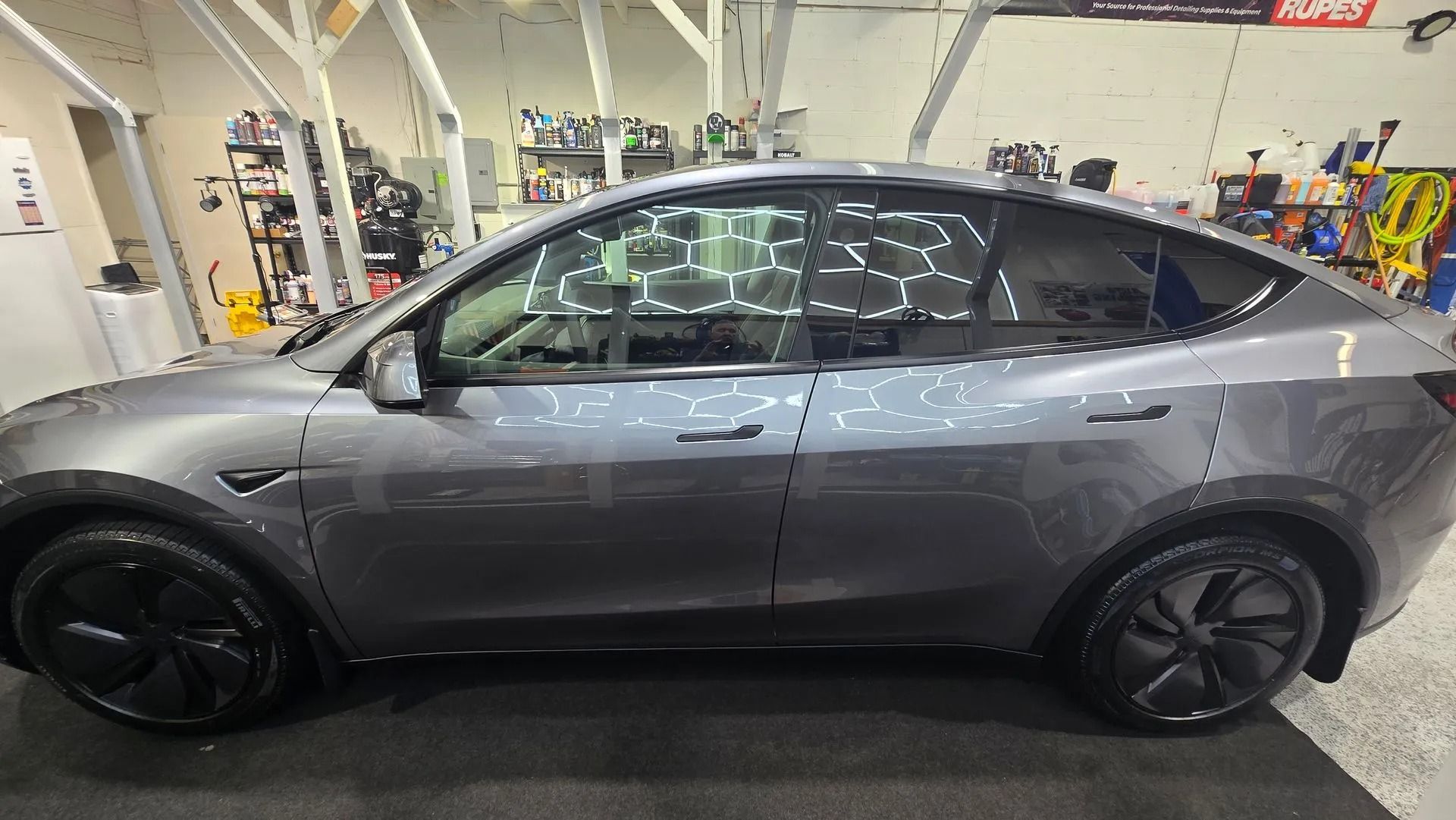 A dark grey Tesla Model Y parked inside a garage, featuring black wheels and tinted windows.
