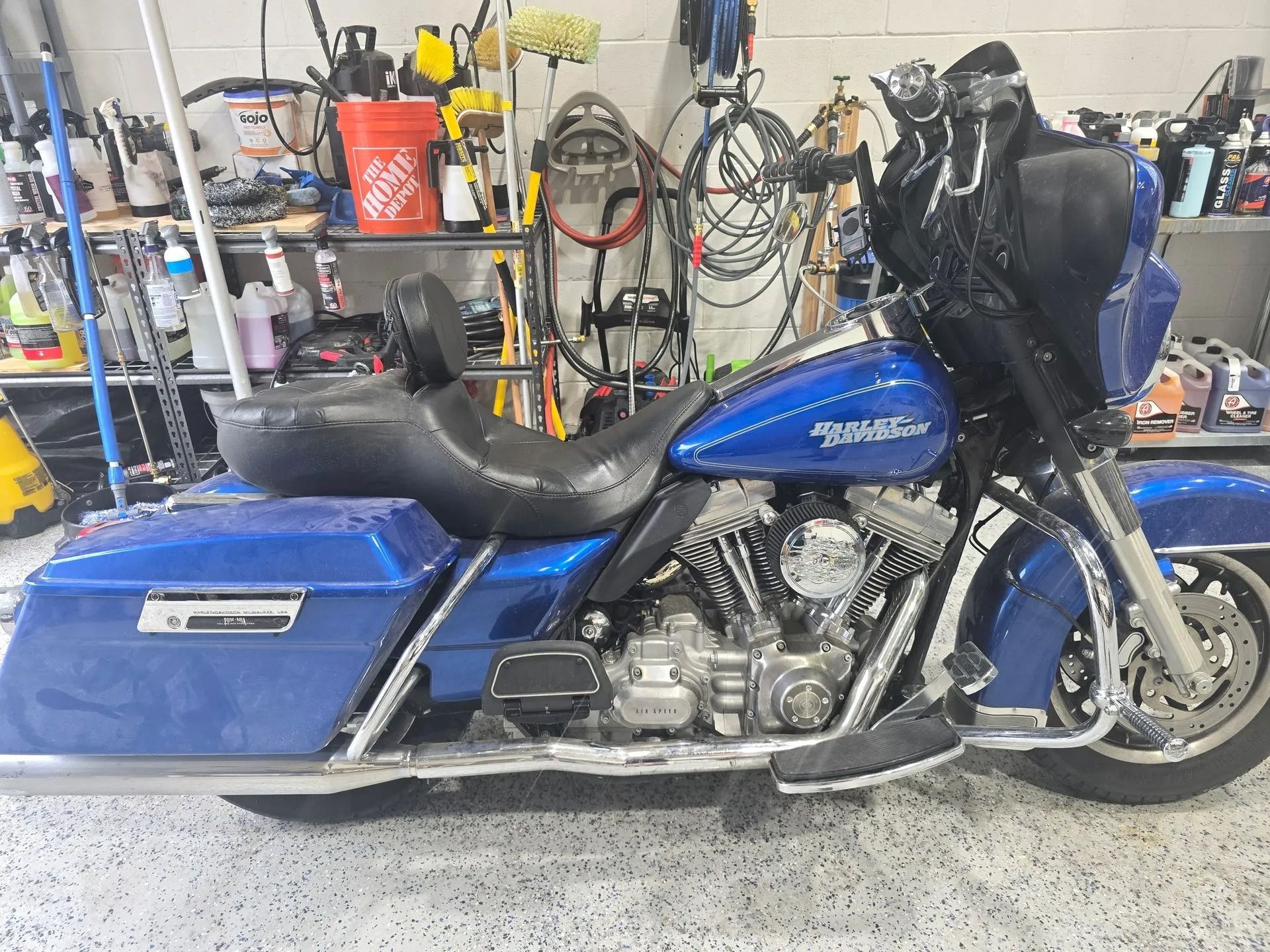 A blue Harley-Davidson motorcycle parked in a cluttered garage with various cleaning supplies and tools in the background.