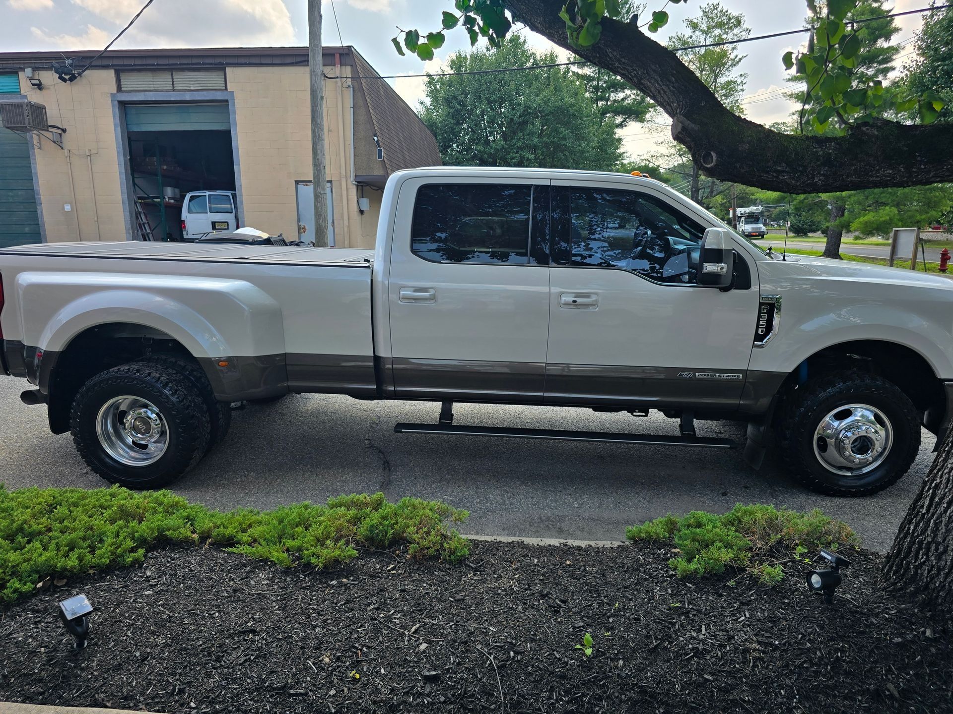 A white and brown dual-rear-wheel Ford pickup truck parked on an asphalt lot beside a building.