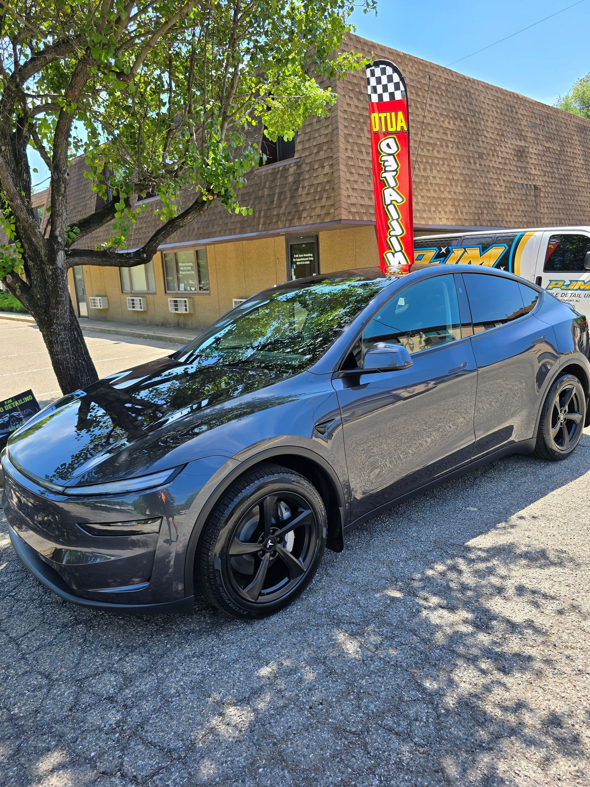 A dark grey Tesla Model Y parked on a gravel lot near a tree and a tall red promotional flag.