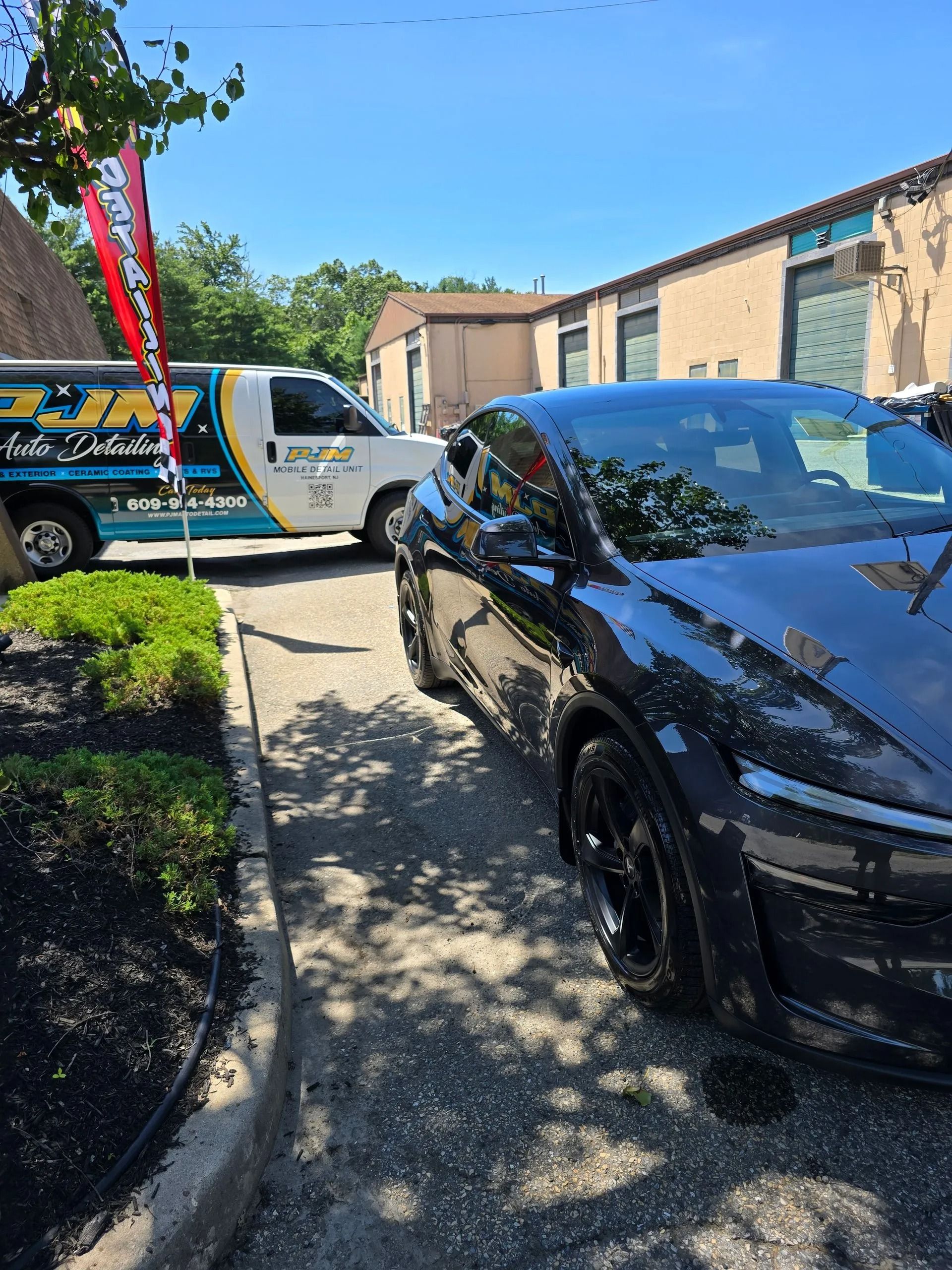 A black Tesla parked next to a white PJM Auto Detailing service van in a paved lot near a tan building.