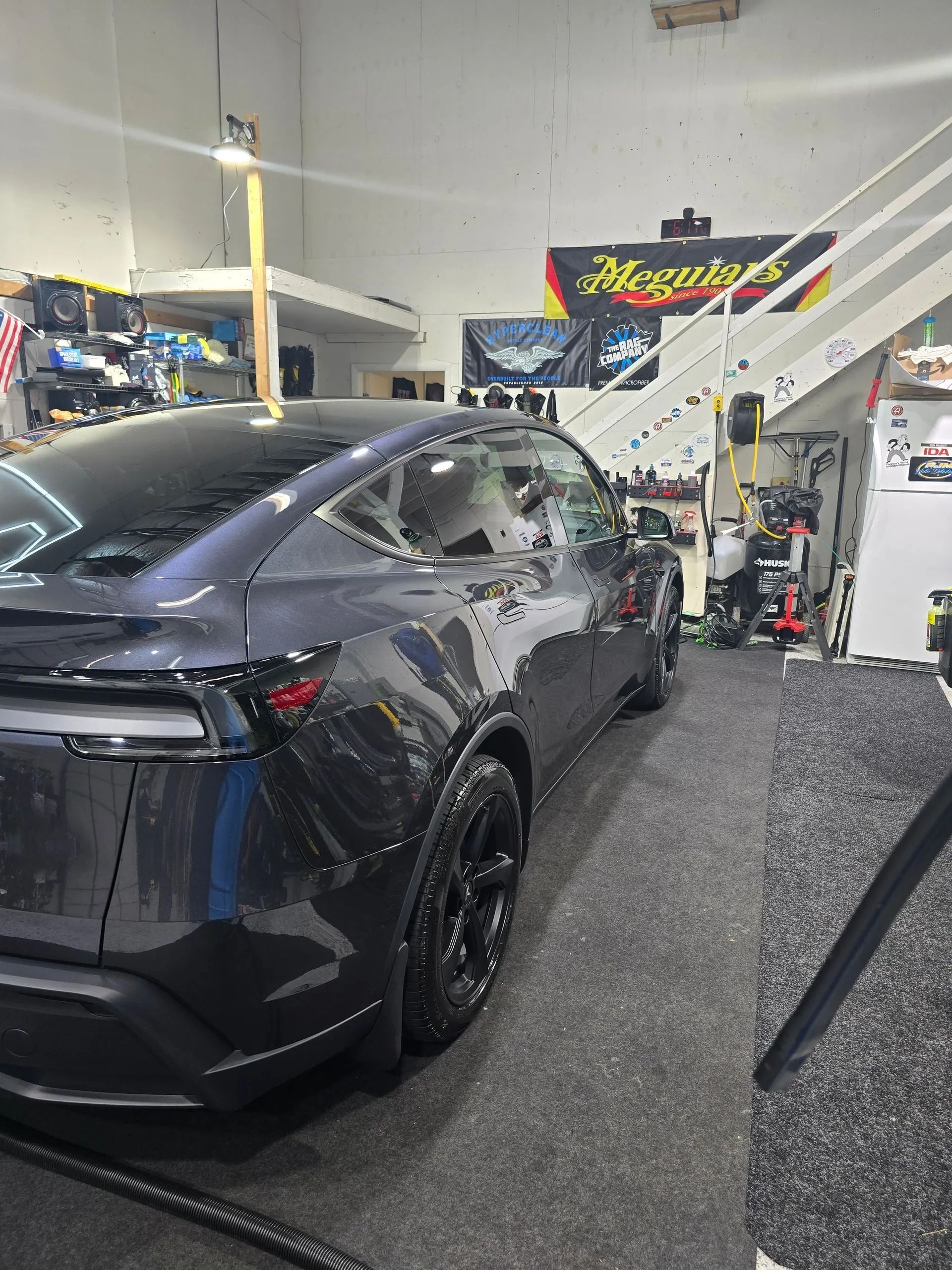 A dark gray Tesla parked inside a garage next to a staircase and detailing supplies, illuminated by overhead lighting.