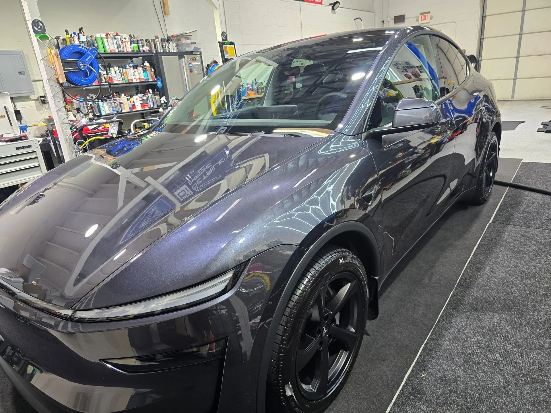 A dark grey Tesla Model Y parked inside a garage workshop with shelves of supplies in the background.