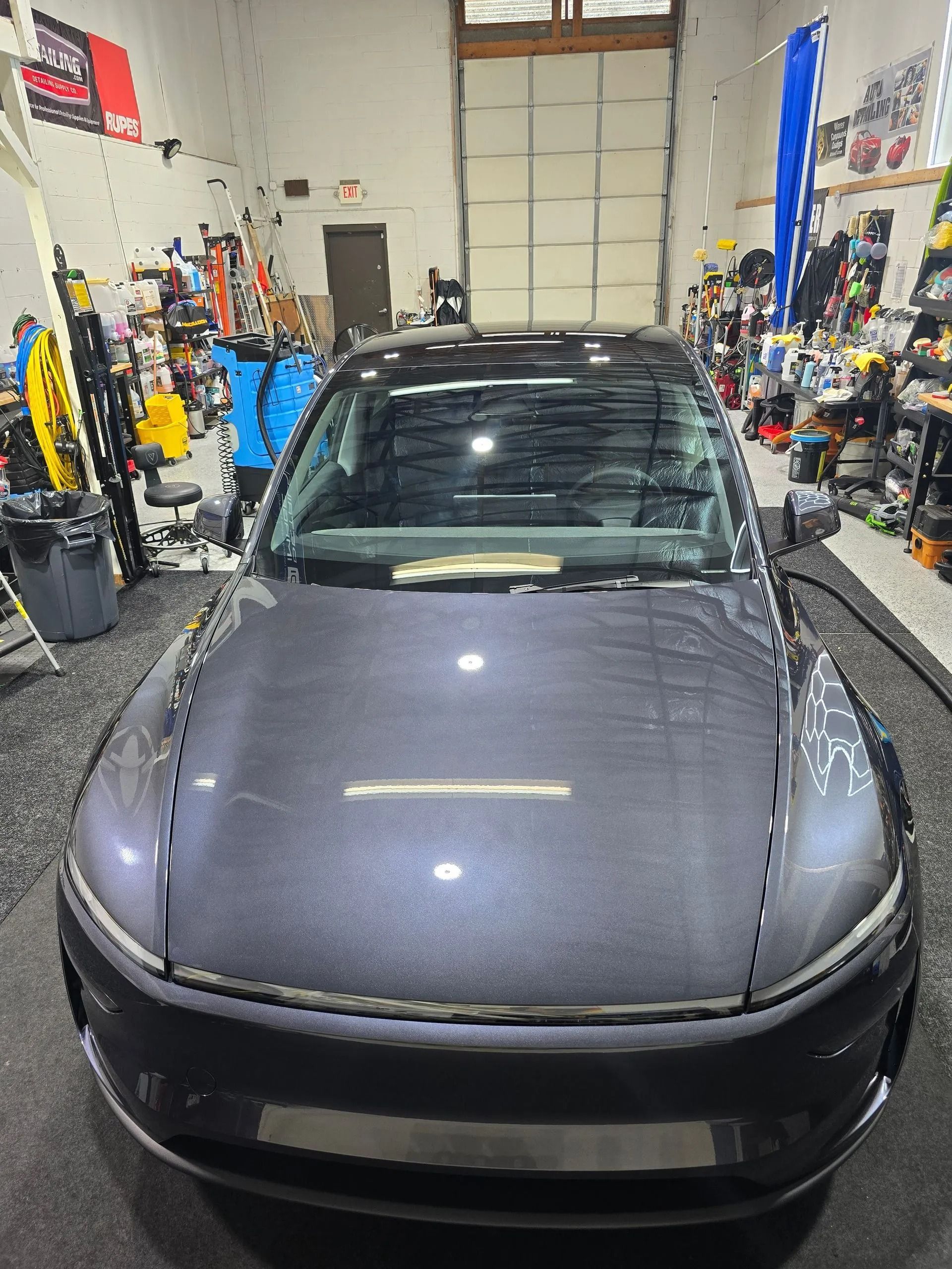 A front-facing view of a dark grey sedan parked in a cluttered automotive garage.