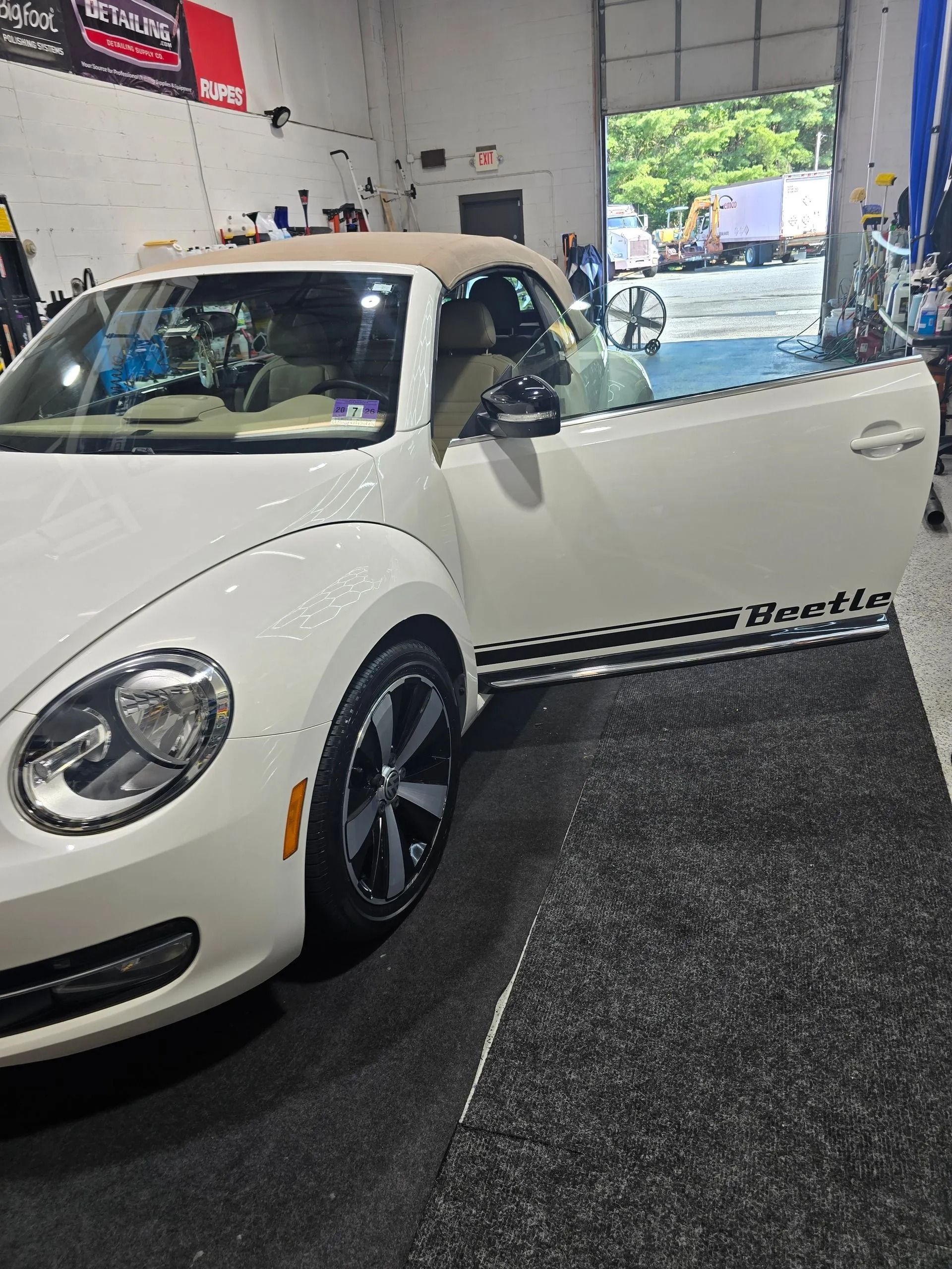 A white Volkswagen Beetle convertible with an open driver’s side door parked inside an auto shop.