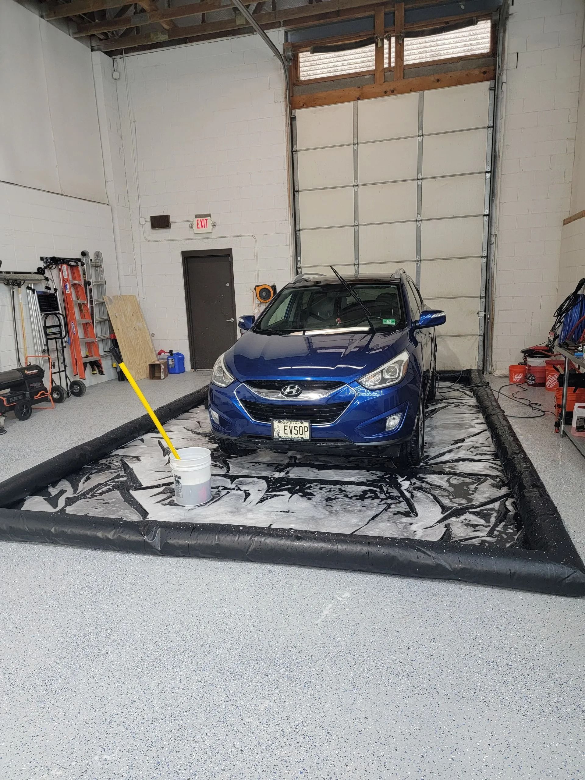 A blue SUV inside a garage, parked within an inflatable containment mat filled with soapy water for washing.